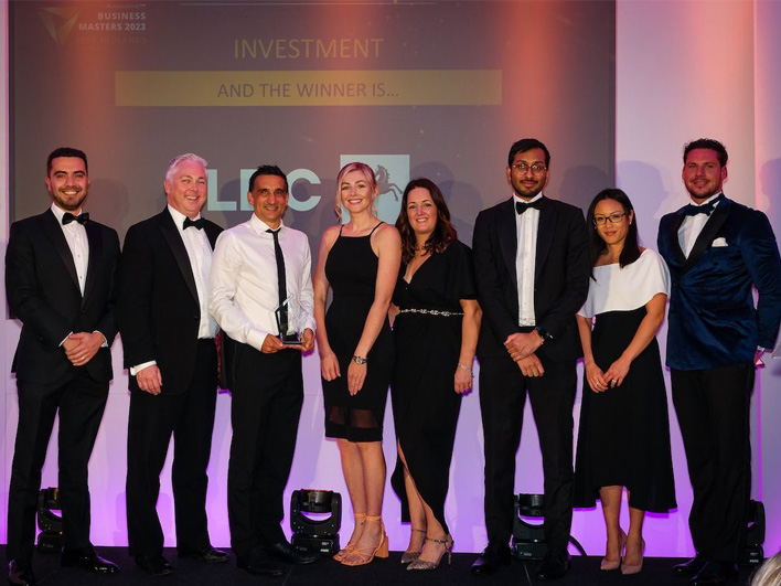 The LDC East Midlands team, dressed in formal evening wear, on stage with a trophy, celebrating a win at the Business Masters 2023 awards, with the announcement screen in the background.