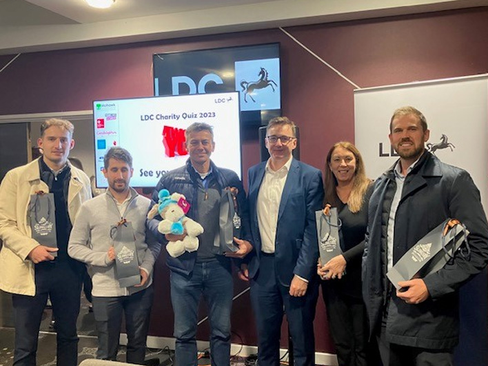 A group of six individuals with prize bags and a stuffed toy, standing in front of a screen displaying 'LDC Charity Quiz 2023' at the event venue.